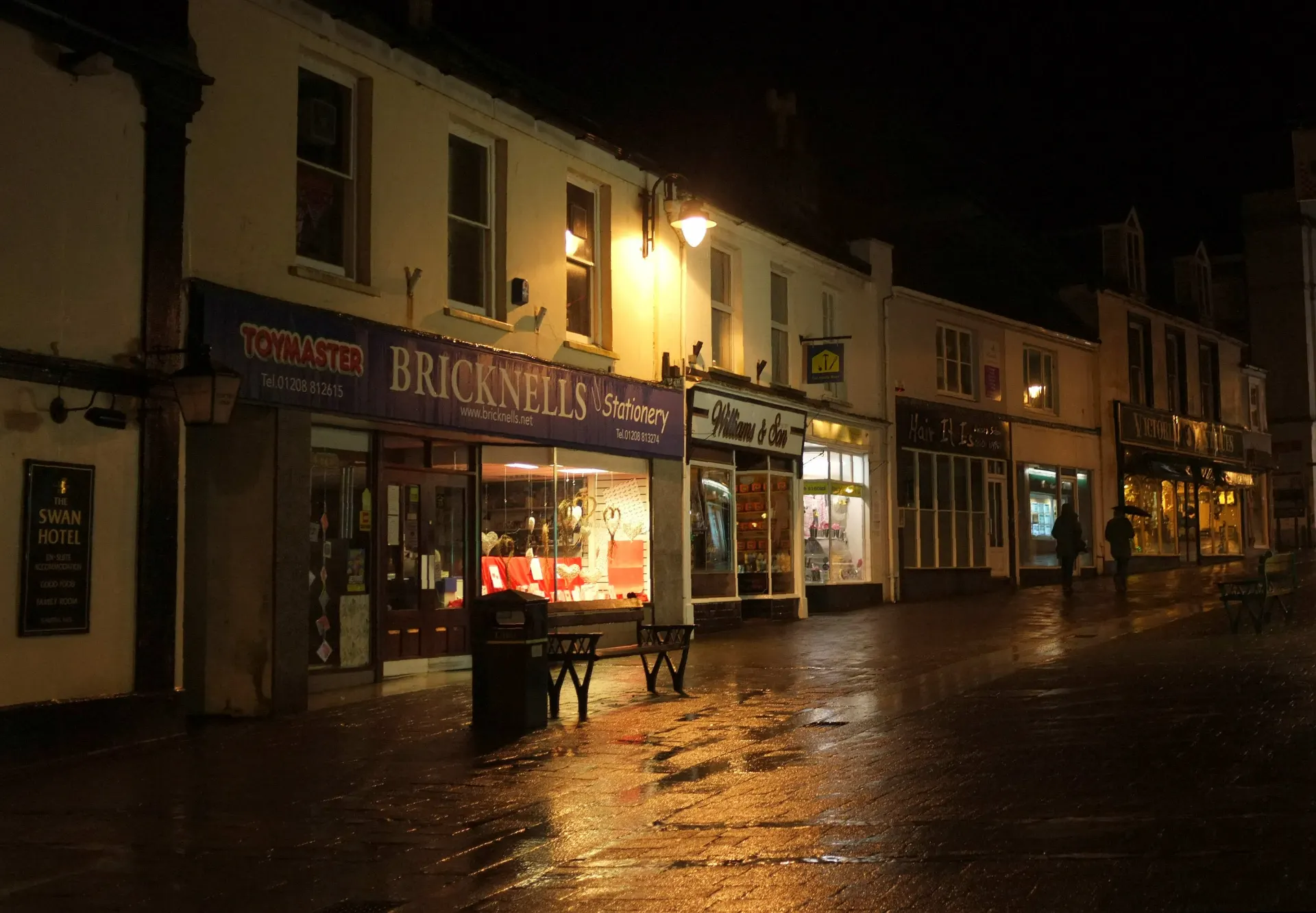 A row of small shops at night, their windows still lit along a wet, empty street